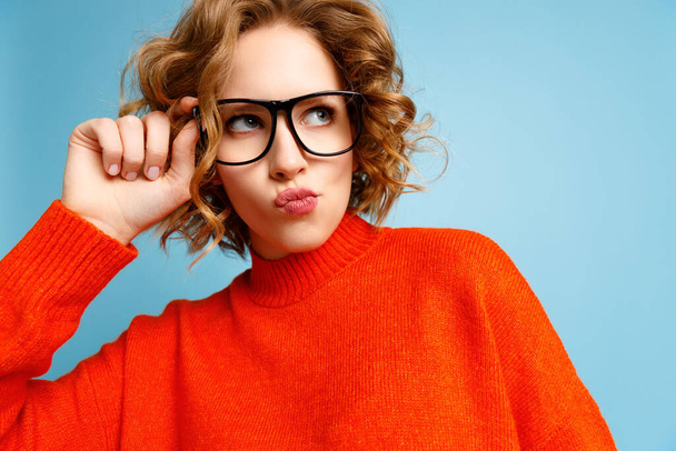 Inquisitive funny female in red sweater adjusting glasses and looking up closely against blue background - Photo, Image