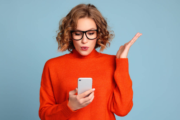 Turuncu süveterli ve gözlüklü genç bir kadın mavi arka planda akıllı telefondan beklenmedik bir mesaj okuyor. - Fotoğraf, Görsel