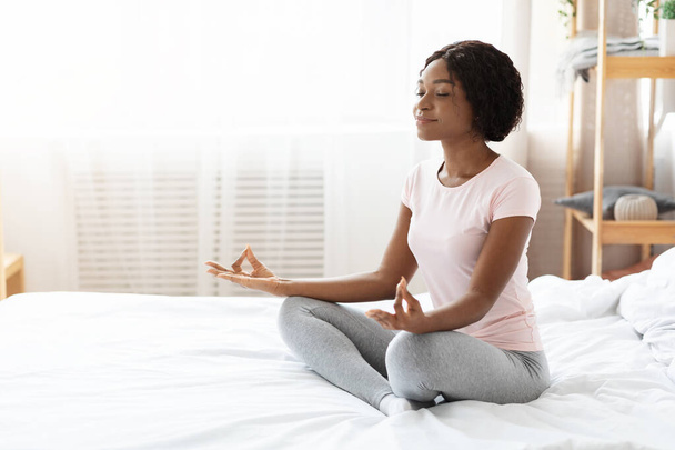 Relaxed black woman sitting on bed and meditating - Φωτογραφία, εικόνα