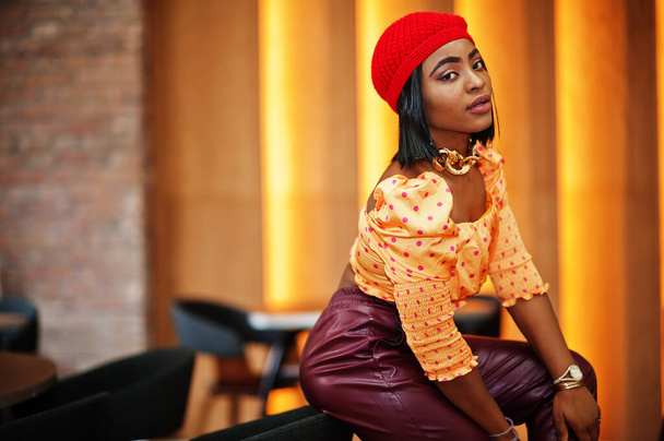 Elegant afro american woman in red french beret, big gold neck chain polka dot blouse and leather pants pose indoor. - Zdjęcie, obraz