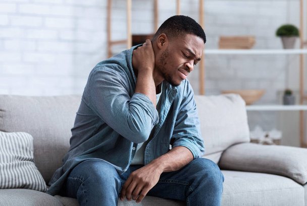 Черный человек страдает от боли в шее, сидя на диване в помещении - Фото, изображение