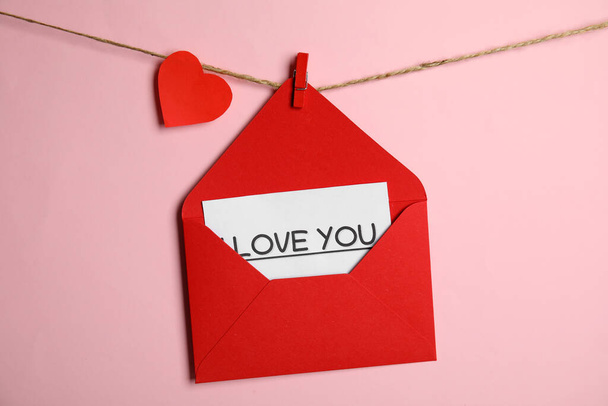 Red envelope with Love You card and paper heart hanging on twine against pink background - Foto, immagini