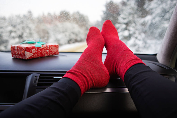 Une fille est assise en chaussettes rouges dans une voiture en hiver, sur fond de forêt hivernale. Concepts voyage, voyage et hiver - Photo, image