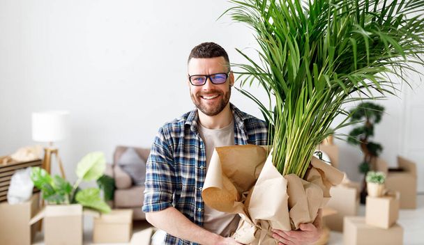 Glad male with potted plant standing in spacious room with carton boxes and looking at camera while moving in new flat - Photo, Image