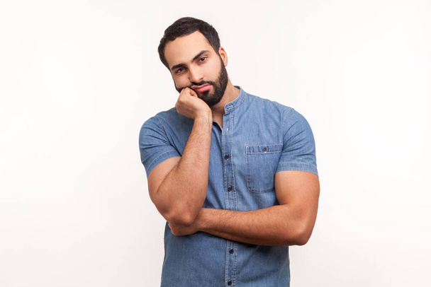 Bored upset man with beard in blue shirt leaning head on hand looking at camera with indifferent look, laziness and apathy, procrastination. Indoor studio shot isolated on white background - Foto, afbeelding