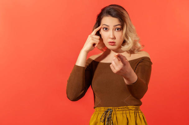 Hey, you are kidding! Portrait of young beautiful woman with surprised face holding finger near head and pointing finger at camera, stupid gesture. Indoor studio shot isolated on red background - Photo, Image