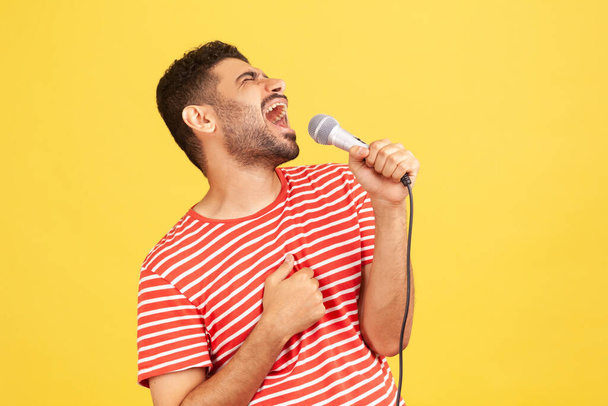 Side view positive bearded man in striped t-shirt loudly singing song holding microphone in hand, having fun resting in karaoke, singer performance. Indoor studio shot isolated on yellow background - Photo, image
