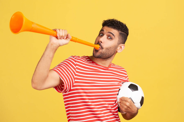 Excited bearded man in red striped t-shirt blowing in horn holding black and white soccer ball, celebrating victory of favourite football team. Indoor studio shot isolated on yellow background - Fotografie, Obrázek