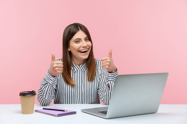Successful brunette woman in striped shirt showing thumbs up sitting at laptop on workplace, looking at camera with smile, satisfied with office work. Indoor studio shot isolated on pink background - 写真・画像