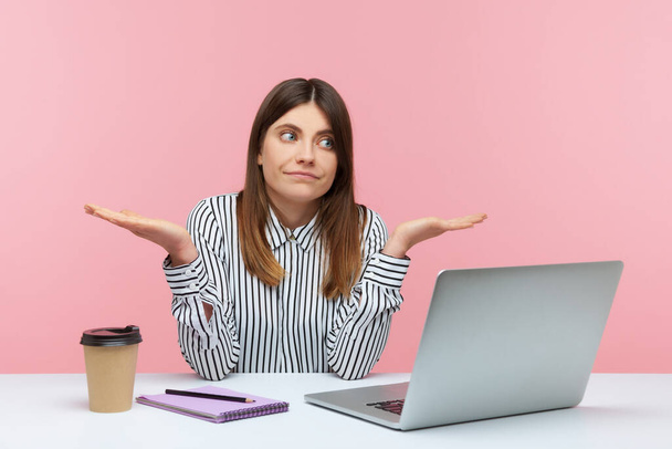 Confused uncertain woman office worker in striped shirt shrugging shoulders sitting on laptop at workplace, not sure about job completion. Indoor studio shot isolated on pink background - Foto, immagini