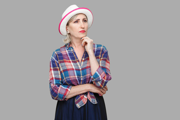 Portrait of serious thoughtful modern stylish mature woman in casual style with white hat standing, touching her chin, query and thinking what to do. indoor studio shot isolated on gray background. - Φωτογραφία, εικόνα