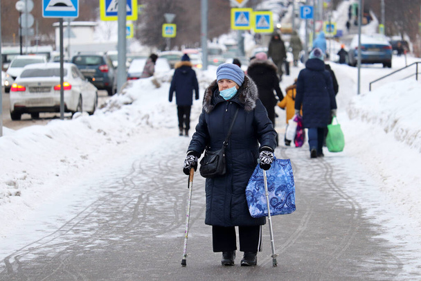 Moscow, Russia - February 2021: Elderly woman in medical mask walking with a canes and bag on a city street on crowd background. Life of old people during covid-19 pandemic - Foto, Imagen