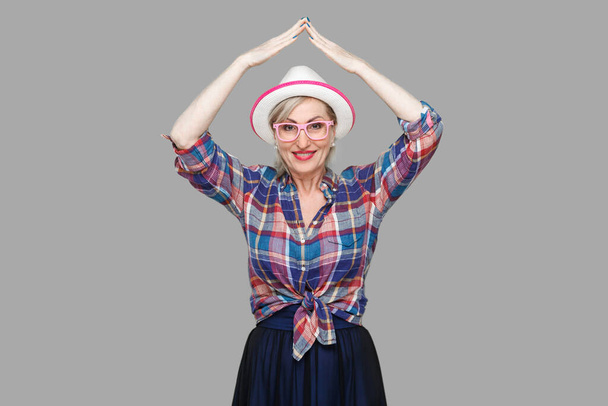 Roof gesture on head. Portrait of happy modern stylish mature woman in casual style with hat standing, holding hand above head, looking with toothy smile. studio shot isolated on gray background. - Photo, image