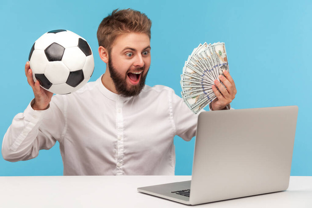 Happy excited man with beard holding fan of dollar cash and soccer ball looking at laptop display, betting and winning jackpot. Indoor studio shot isolated on blue background - Φωτογραφία, εικόνα