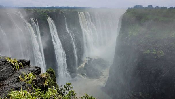 Powerful streams of water rush down from the rocks. In a deep gorge, there is fog from water splashes. In the foreground, wet stones and green grass. Victoria Falls. Zimbabwe. - Foto, afbeelding