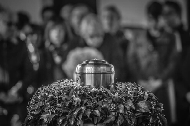 Une urne en métal avec des cendres d'une personne décédée lors d'un enterrement, avec des gens en deuil en arrière-plan sur un service commémoratif. Triste moment de deuil à la fin d'une vie. Dernier adieu à une personne dans une urne. - Photo, image