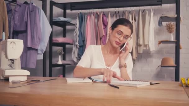 Seamstress falando no smartphone perto de notebooks e máquina de costura  - Filmagem, Vídeo
