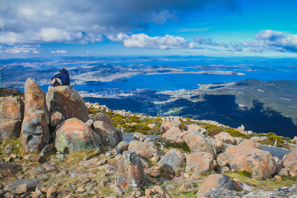 Ένα ζευγάρι κάθεται σε ένα σχηματισμό βράχων και απολαμβάνοντας τη θέα πάνω από Hobart από την κορυφή του όρους Ουέλινγκτον, Hobart, Αυστραλία. - Φωτογραφία, εικόνα