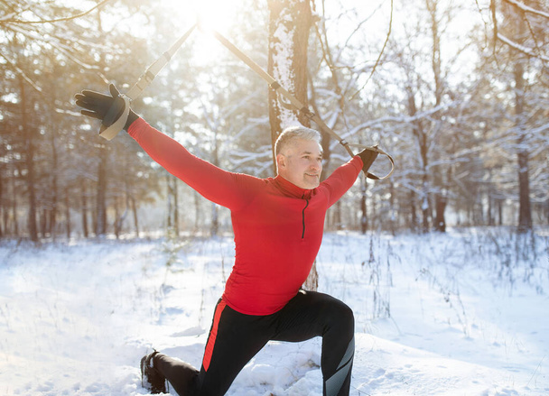 Concepto de entrenamiento en suspensión TRX. Hombre mayor atlético haciendo ejercicio con equipo deportivo en el día de invierno nevado en el parque - Foto, imagen