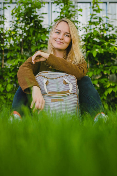 Hipster young blonde woman with septum piercing sitting on a grass in park, posing with a fashionable backpack. Leisure activity and relax after school for a young woman. - Photo, Image