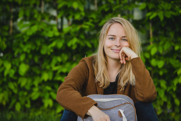 Young blonde hipster girl seductevly looking and smiling at a camera while supporting her chin with one arm and other resting on a fashionable striped backpack. Green bush in the background. - Photo, Image