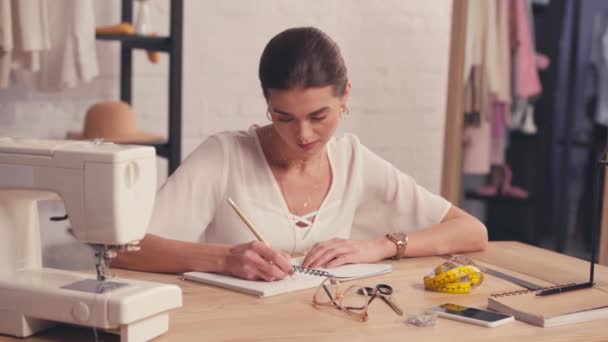 Designer schrijven op notebook in de buurt van naaimachine en smartphone  - Video