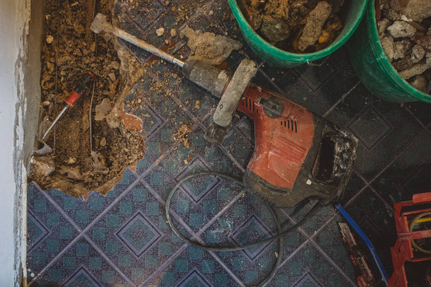 Marteau de démolition électrique rouge sur le sol, tout en rénovant un vieux tuyau d'eau dans le sol. Site de construction avec outils, vieux matériaux et gants est vu. - Photo, image