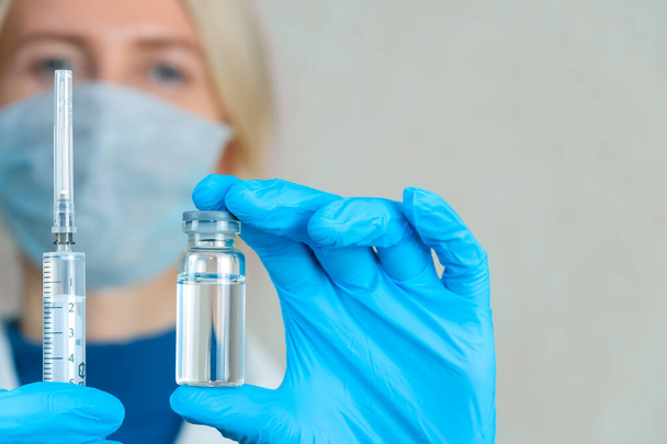 Médica em um casaco branco, máscara e luvas azuis segura uma seringa em uma mão, e na outra um frasco de medicamento close-up, copiar o espaço. Banner. Conceito de medicamento, medicamentos e vacinação - Foto, Imagem
