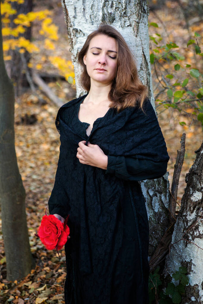 portrait of sad brunette woman in black dress holding red flower in forest - Foto, afbeelding
