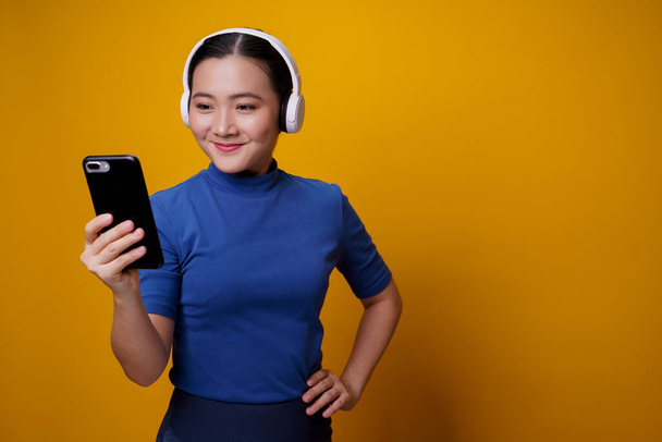 黄色の背景に隔離されたスマートフォンから音楽を聴くヘッドフォンを持つアジアの女性. - 写真・画像
