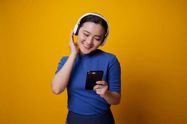 Asian woman with headphones listening to music from smart phone isolated on yellow background. - Zdjęcie, obraz