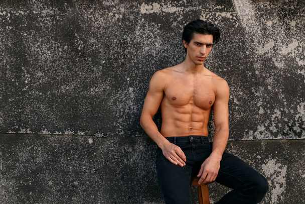 Outside portrait of a hot fit young man with muscular body and bare torso showing six pack abs posing at old black wall. Copy space. - Photo, Image