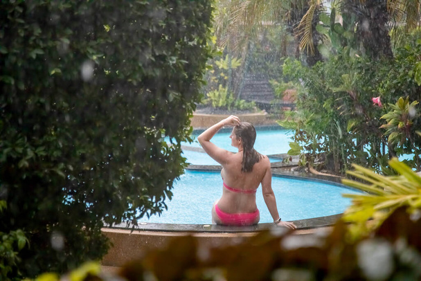 ένα κορίτσι με μπικίνι κάθεται στο πλάι μιας εξωτερικής πισίνας σε έναν τροπικό κήπο στη βροχή. - Φωτογραφία, εικόνα