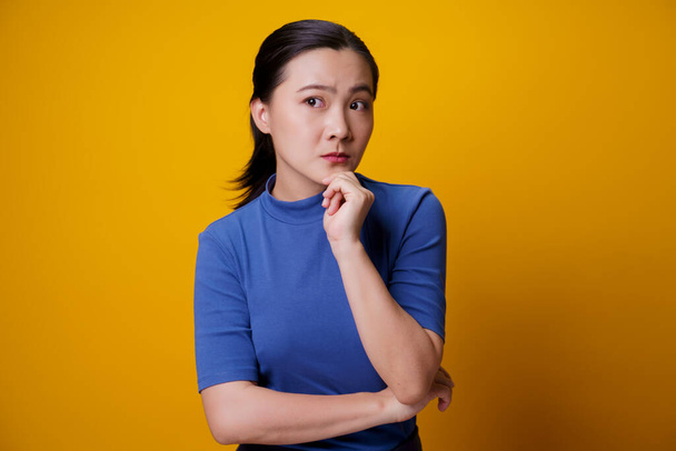 Verwirrte asiatische Frau denkt und steht isoliert auf gelbem Hintergrund. - Foto, Bild