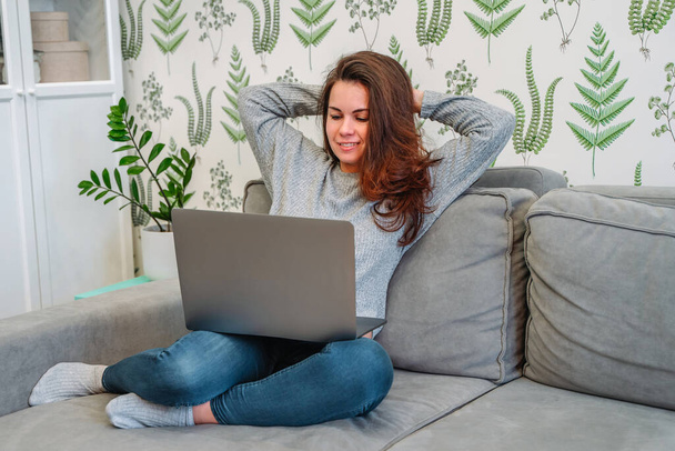 Hymyilevä nuori nainen freelancer istuu sohvalla nauttien etätyöstä kotoa. Onnellinen nainen opiskelija opiskelee online kursseja sisätiloissa - Valokuva, kuva