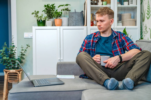 A young man holds a laptop on his lap while sitting on the couch and work remotely from home. Distance learning online-education and work. - Photo, Image