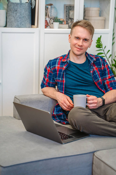 Молодой человек держит ноутбук на коленях, сидя на диване и работая удаленно от дома. Дистанционное обучение онлайн-образование и работа. - Фото, изображение