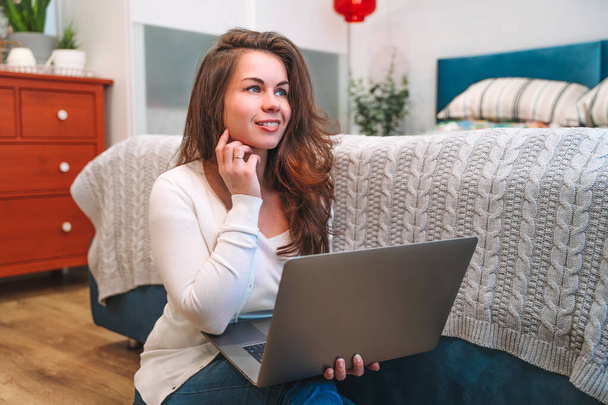 Egy modern fiatal nő laptopot használ, ami a meleg padlón ül az ágy mellett a hálószobában. Távmunka otthonról - Fotó, kép