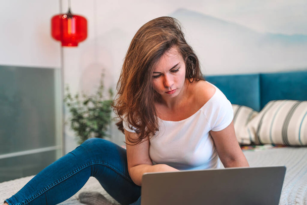 Una joven usa una computadora portátil mientras está acostada en la cama. El concepto de la oficina en casa y la educación en línea. - Foto, imagen