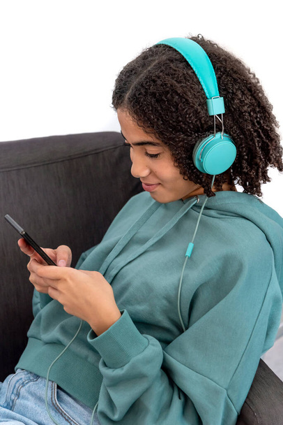 Черный подросток в повседневной одежде просматривает смартфон и слушает музыку, отдыхая в кресле дома - Фото, изображение