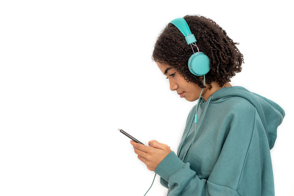Adolescente afroamericano con auriculares turquesa y sudadera con capucha usando smartphone y escuchando música - Foto, Imagen