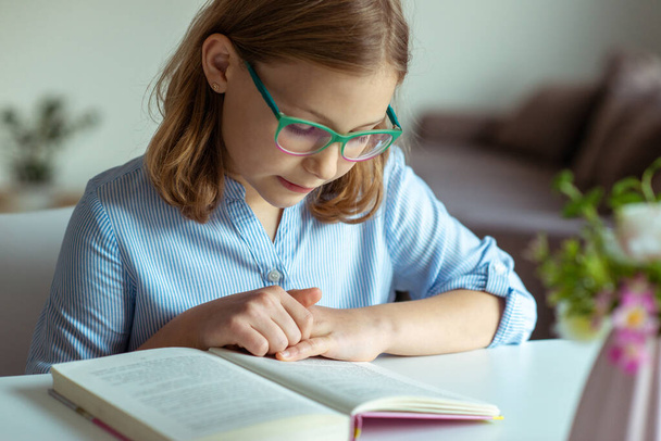 Happy pretty teen girl reading book at desk at home during pandemic distance learning  - 写真・画像