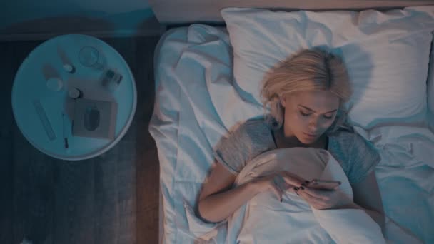 ylhäältä näkymä nainen käyttää älypuhelinta, kun makaa sängyssä lähellä yöpöytä - Materiaali, video