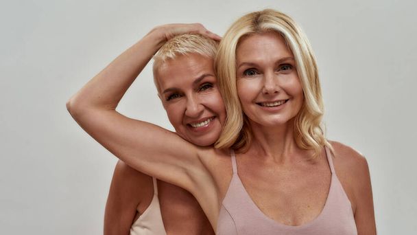 Studio portrait of two happy blonde women smiling at camera, standing together, posing isolated over gray background - 写真・画像