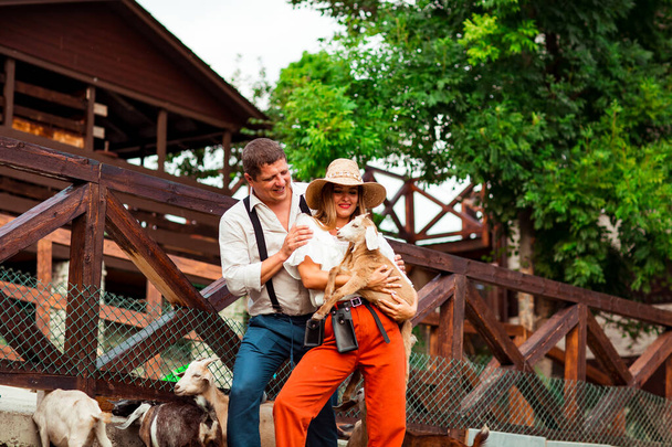een gelukkig gezin, een man en een vrouw met een hoed, ontspannen en lachen op een boerderij, omringd door geiten, op een zonnige zomerdag - Foto, afbeelding