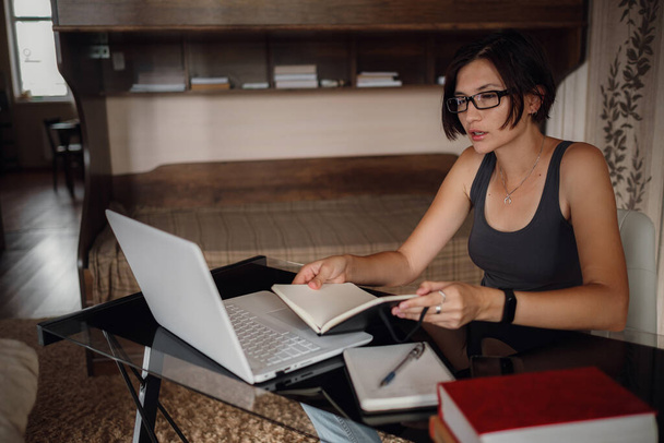 Young asian female student in glasses using laptop, read a book. Cozy office workplace, remote work, E learning concept. - Photo, Image