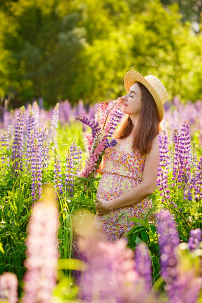 Üzerinde çiçekli bir taç ve büyük mavi lupinli hamile bir kadın çiçek açan lupin tarlasında oturuyor. - Fotoğraf, Görsel