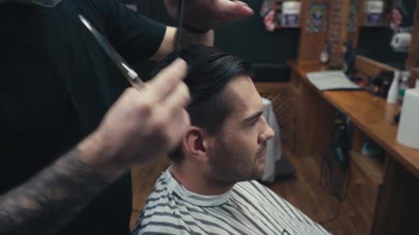 Tattooed barber cutting hair of man in cape in barbershop  - Filmagem, Vídeo