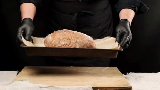 Hombre del chef en uniforme negro, poner una bandeja de metal para hornear en la mesa con pan redondo al horno, fondo negro - Imágenes, Vídeo