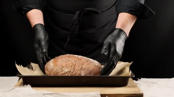 hombre chef en uniformes negros y guantes de látex negro sostiene pan redondo horneado sobre una bandeja de metal para hornear - Imágenes, Vídeo
